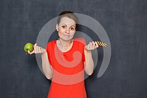 Portrait of doubting girl holding apple and cookie in hands