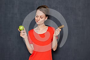 Portrait of doubting girl making choice between apple and cookie