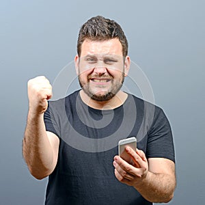 Portrait of ecstatic man holding cell phone and celebrating with closed fist against gray background