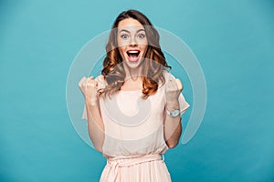 Portrait of ecstatic woman 20s wearing dress screaming and clenching fist like rejoicing victory or triumph, isolated over blue b