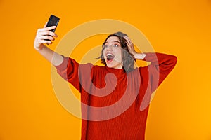 Portrait of ecstatic woman holding and taking selfie photo on cell phone while standing isolated over yellow background