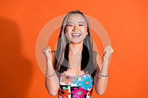 Portrait of ecstatic woman with straight hairdo dressed print tank clenching fists cream win lottery isolated on orange