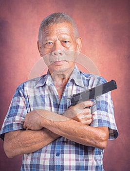 Portrait of an elderly Asian man holding a short gun and looking at the camera while standing with a brown background. Space for