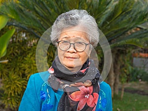 Portrait of an elderly Asian woman wearing glasses, smiling, and looking at the camera while standing in a garden