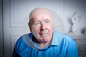 Portrait of an elderly bald man in a blue shirt in the room. A poor pensioner lives alone