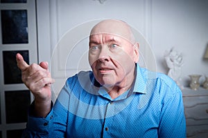 Portrait of an elderly bald man in a blue shirt in the room. A poor pensioner lives alone