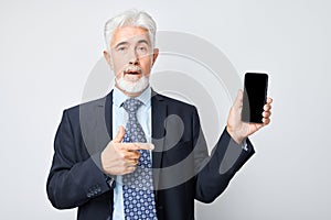 Portrait of elderly businessman in suit showing blank mobile phone screen with excited face. Person with smartphone