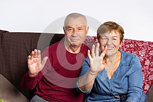 Portrait of an elderly couple sitting on the couch and waving hands in front of the camera