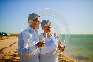 Portrait of elderly couple standing together with glasses of wine and embracing on the beach.