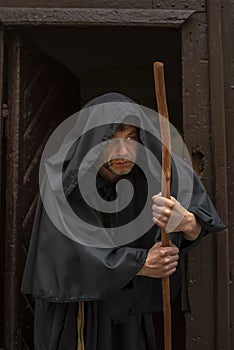 Portrait of an elderly hunched monk 45-50 years old with a beard in a black robe with a staff coming out of the door of a monaster