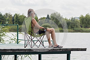 Portrait of an elderly man aged 60-65 with a beard sitting in a tourist chair on a wooden platform against the backdrop of the lak