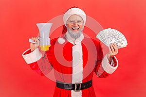 Portrait of elderly man with gray beard wearing santa claus costume holding fan of banknotes and megaphones, looking at camera