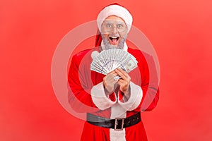 Portrait of elderly man with gray beard wearing santa claus costume holding fan of dollars, looking