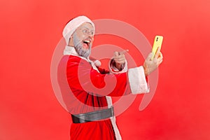 Portrait of elderly man with gray beard wearing santa claus costume standing with smart phone