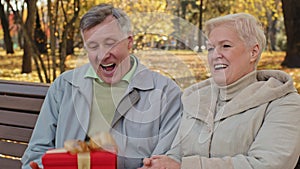 Portrait elderly man rejoices gift surprise mature grandpa throwing red box feel happy married couple sitting on bench