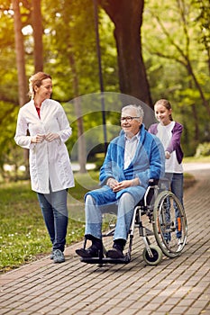 portrait of elderly man on wheelchair with nurse and granddaughter outdoor.