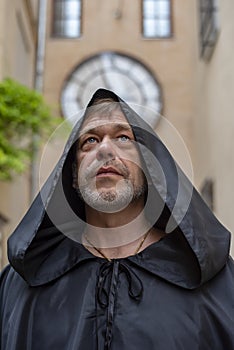 Portrait of an elderly monk 45-50 years old with a beard in a black cassock against the background of old buildings and clocks.