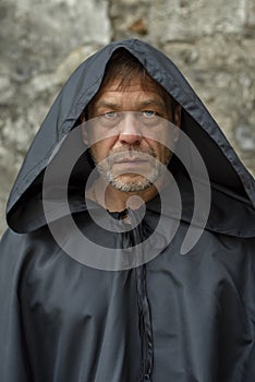 Portrait of an elderly monk 45-50 years old with a beard in a black robe with a staff on the background of a textured wall, lookin