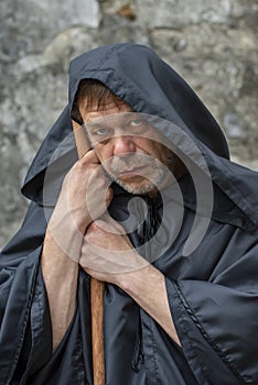 Portrait of an elderly monk 45-50 years old with a beard in a black robe with a staff on the background of a textured wall, lookin