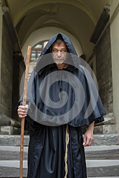 Portrait of an elderly monk 45-50 years old with a beard in a black robe with a staff, standing under the arches of a monastery or