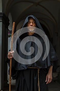 Portrait of an elderly monk 45-50 years old with a beard in a black robe with a staff, standing under the arches of a monastery or