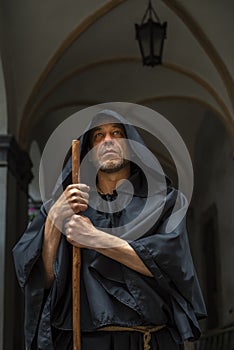 Portrait of an elderly monk 45-50 years old with a beard in a black robe with a staff, standing under the arches of a monastery or