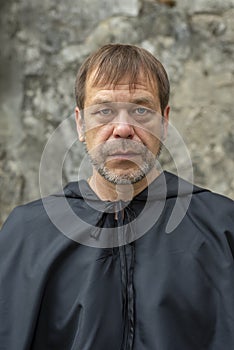 Portrait of an elderly monk 45-50 years old with a beard in a black robe on the background of a textured wall, looking directly in
