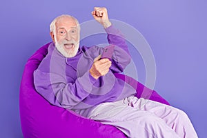 Portrait of elderly retired trendy cheerful lucky grey-haired man sitting in chair rejoice isolated over bright violet