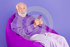 Portrait of elderly retired trendy cheery grey-haired man sitting in bag chair using device isolated over bright violet