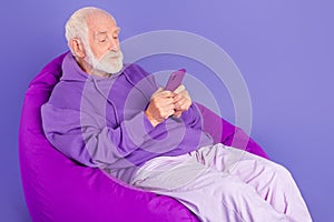 Portrait of elderly retired trendy grey-haired man sitting in chair using device typing isolated over bright violet