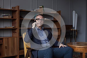 Portrait of elderly successful businessman in blue suit and glasses talking on mobile phone on grey office wall background