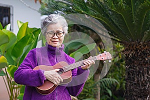 Portrait of an elderly woman playing the ukulele while standing in the garden. Relaxing by singing and play small guitar happy