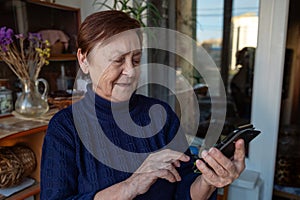 Portrait of an elderly woman using a smartphone. Elderly woman near window at home using mobile phone