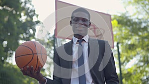 Portrait of elegant African American businessman posing with ball on basketball court. Middle shot of confident young