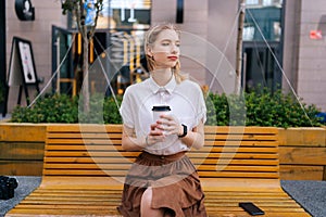 Portrait of elegant blonde young woman sitting on bench holding paper cup of takeaway coffee pensive looking away.