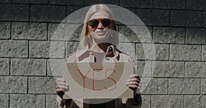 Portrait of elegant businesswoman holding YES sign standing outdoors against brick wall background