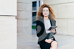 Portrait of elegant lady dressed in white blouse, black jacket and skirt, holding tablet and pocket book while sitting near office