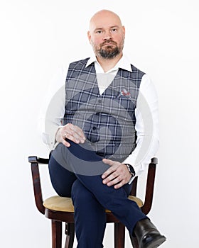 Portrait of elegant middle-aged businessman in grey vest, blue jeans, white shirt sitting on chair with legs crossed.