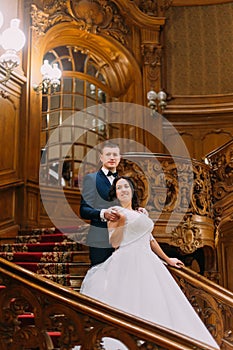 Portrait of elegant newlywed pair posing on stairs in rich interior at old classic mansion