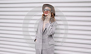 Portrait elegant smiling woman calling on smartphone wearing gray coat, round hat looking away over white wall
