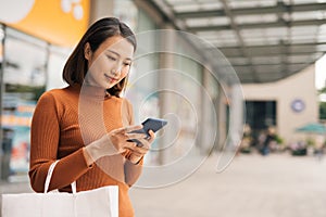 Portrait of elegant young Asian woman holding shopping bags and using smartphone on the go while leaving mall