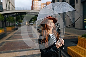 Portrait of elegant young woman in fashion hat standing on beautiful city street with transparent umbrella enjoying