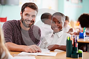 Portrait Of Elementary School Teacher Giving Male Pupil Wearing Uniform One To One Support In Class