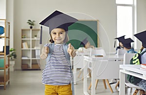 Portrait of happy school girl in square academic cap smiling and giving thumbs up