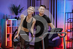 Portrait of embracing young couple doing cardio training on exercise machines at evening time.
