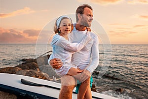 Portrait of embrasing father and daughter posing at background with sup board and sunset ocean. Family summer vacations