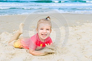 Portrait of emotional adorable toddler girl in sun protecting swimming suit lying on the sand on the beach and looking at camera.