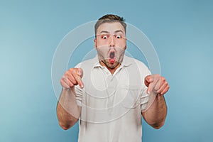 Portrait of emotional crazy man in white t-shirt on blue background looks with shocked face and points thumbs down