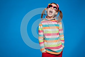 Portrait of an emotional girl of five years old in a striped sweater and in children`s glasses on a blue background