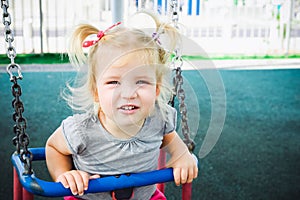 Portrait of emotional little child blond girl looking at camera and having fun on a swing outdoor.Baby Summer playground. Selectiv